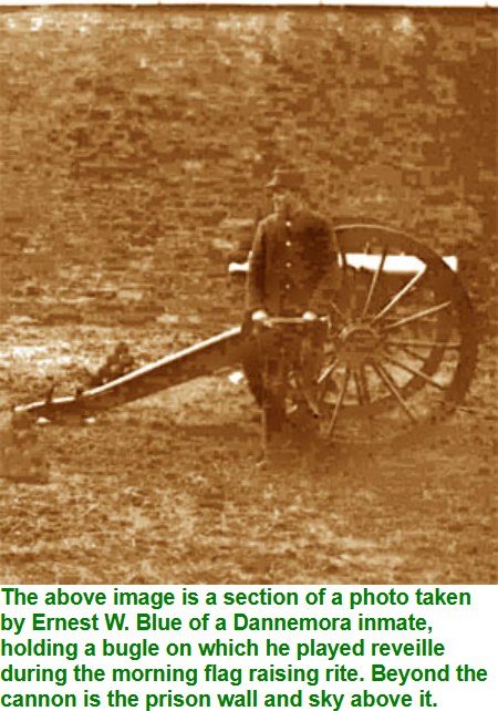 Sepia photograph of a uniformed man standing beside a field cannon in front of a high prison wall, holding a bugle.
