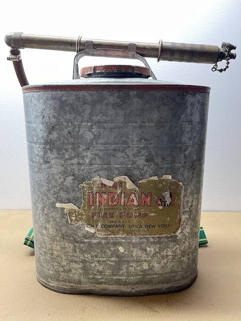A galvanised Indian fire pump: an oval five-gallon back tank with a red rim, a carrying handle and a long brass trombone hand pump, the paper label worn.